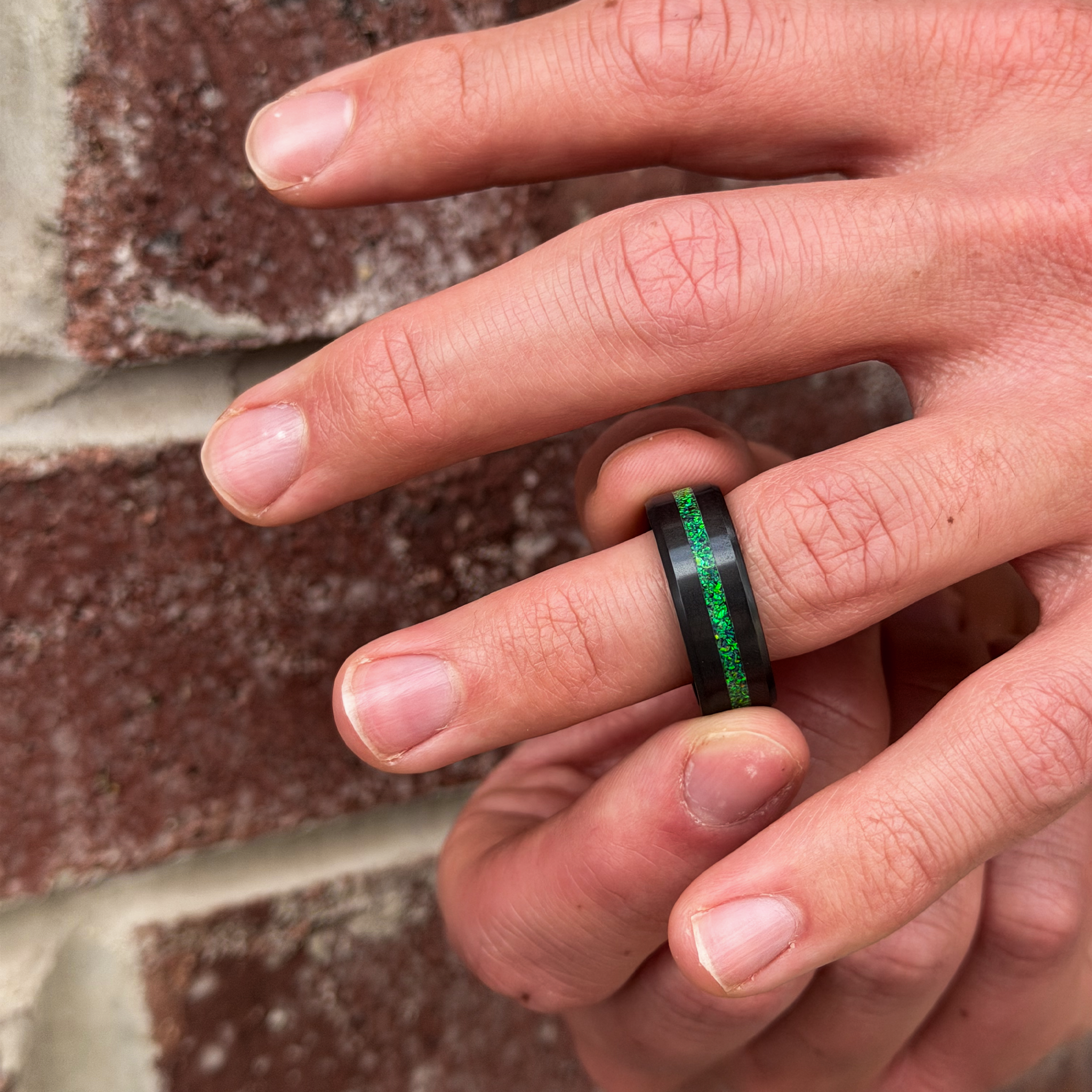 A solid black diamond ring with a green opal is worn on a finger with a brick wall in the background.