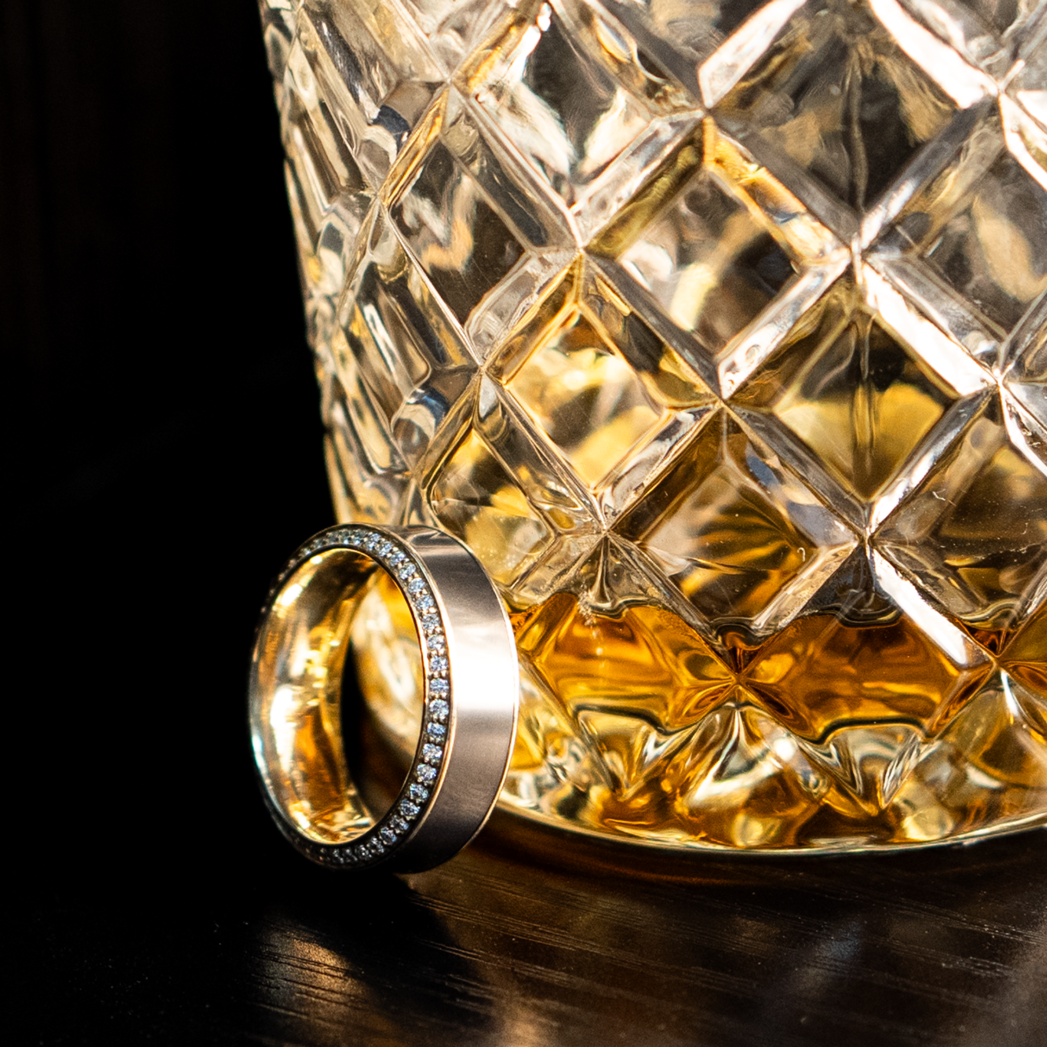 HELIOS ring – 8mm black diamond men’s ring with a yellow gold band, black diamond inlay, and white diamond insets. Resting beside a whiskey glass on a wooden table, highlighting its refined luxury and masculine edge.