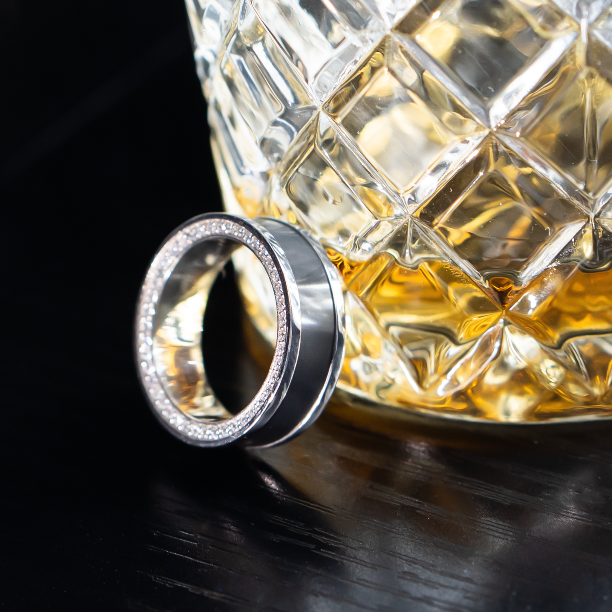 ZEUS ring – 7mm black diamond men’s ring with a platinum band, black diamond inlay, and white diamond insets. Resting beside a whiskey glass on a wooden table, highlighting its refined luxury and masculine edge.