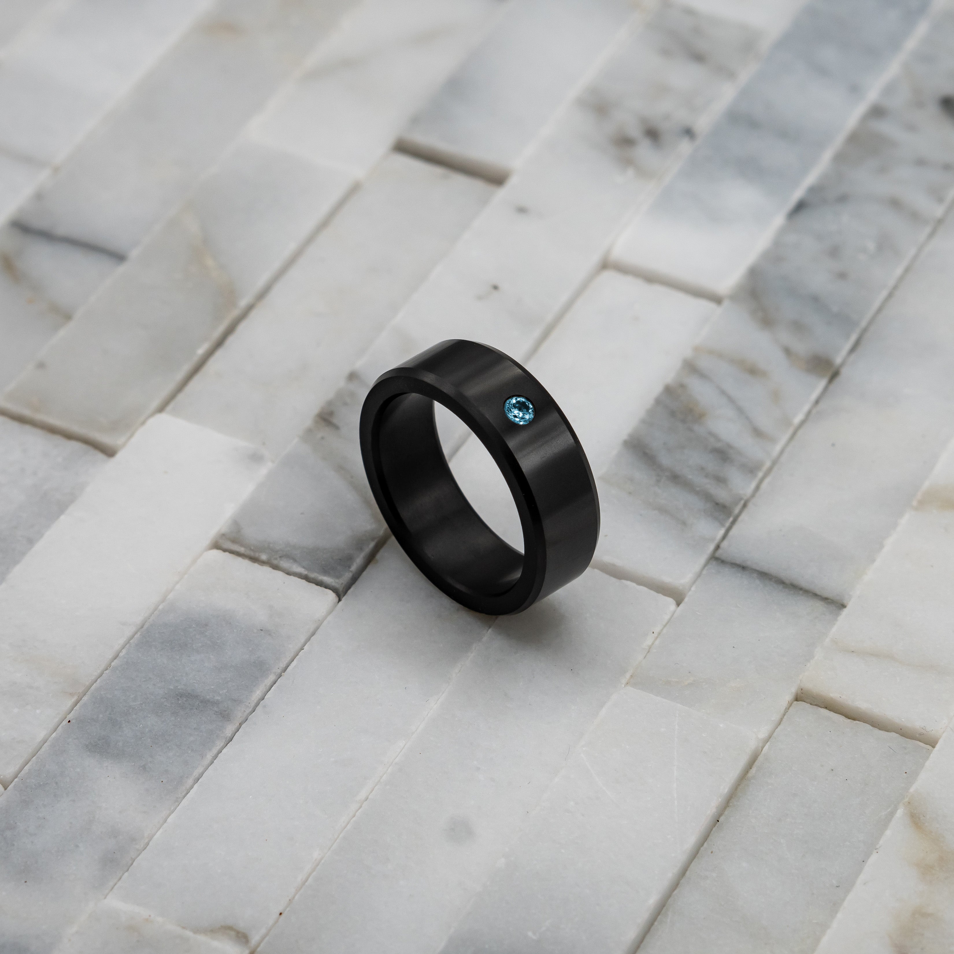 Ring shot #2 of our Elysium ARES Men's Black Diamond & Blue Diamond Inset resting on a marble surface | ElysiumBlack.com | Men’s Blue Diamond Wedding Rings